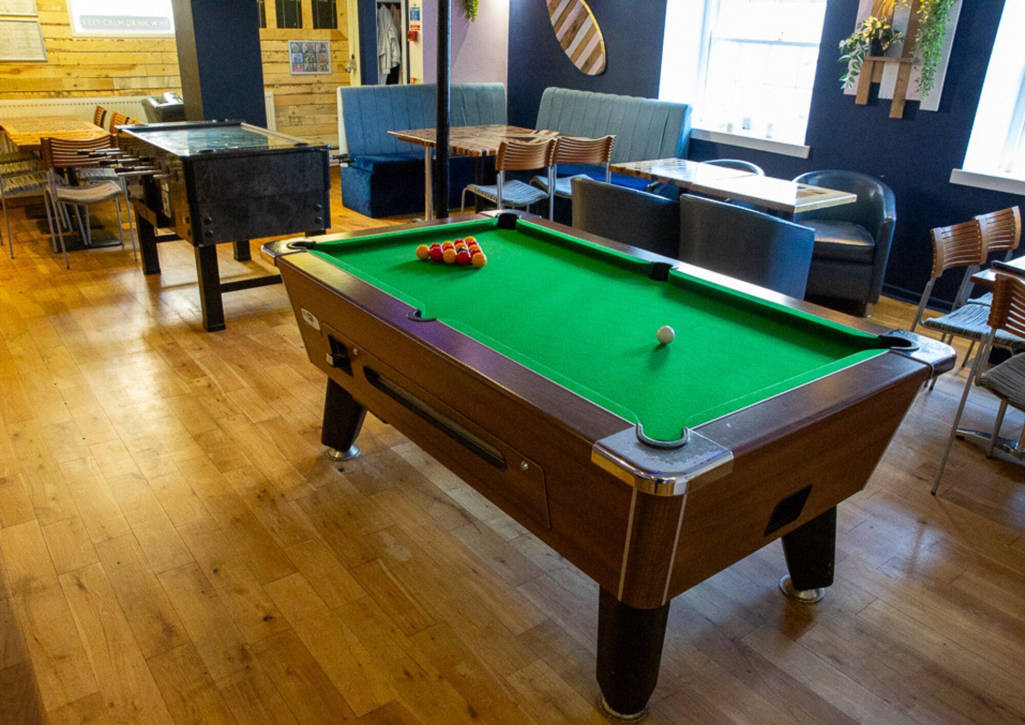 A pool table in a room with wooden flooring, seating arrangements, and a window with plants.