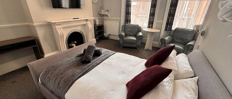 A hotel room with a large bed, two armchairs, a fireplace, and a television.