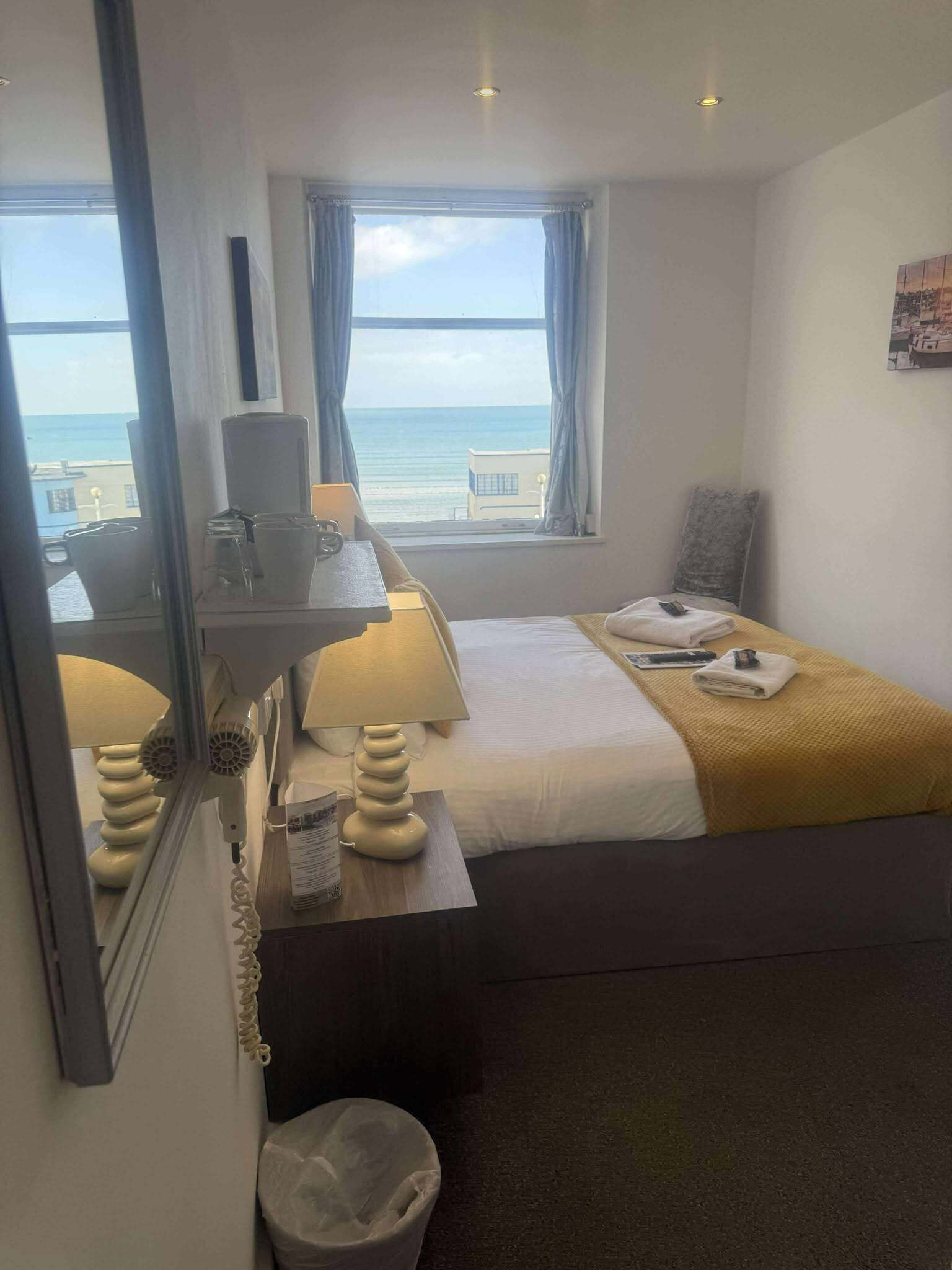 A hotel room with a bed, a bedside table with a lamp, a window with a view of the sea, and a small trash bin.