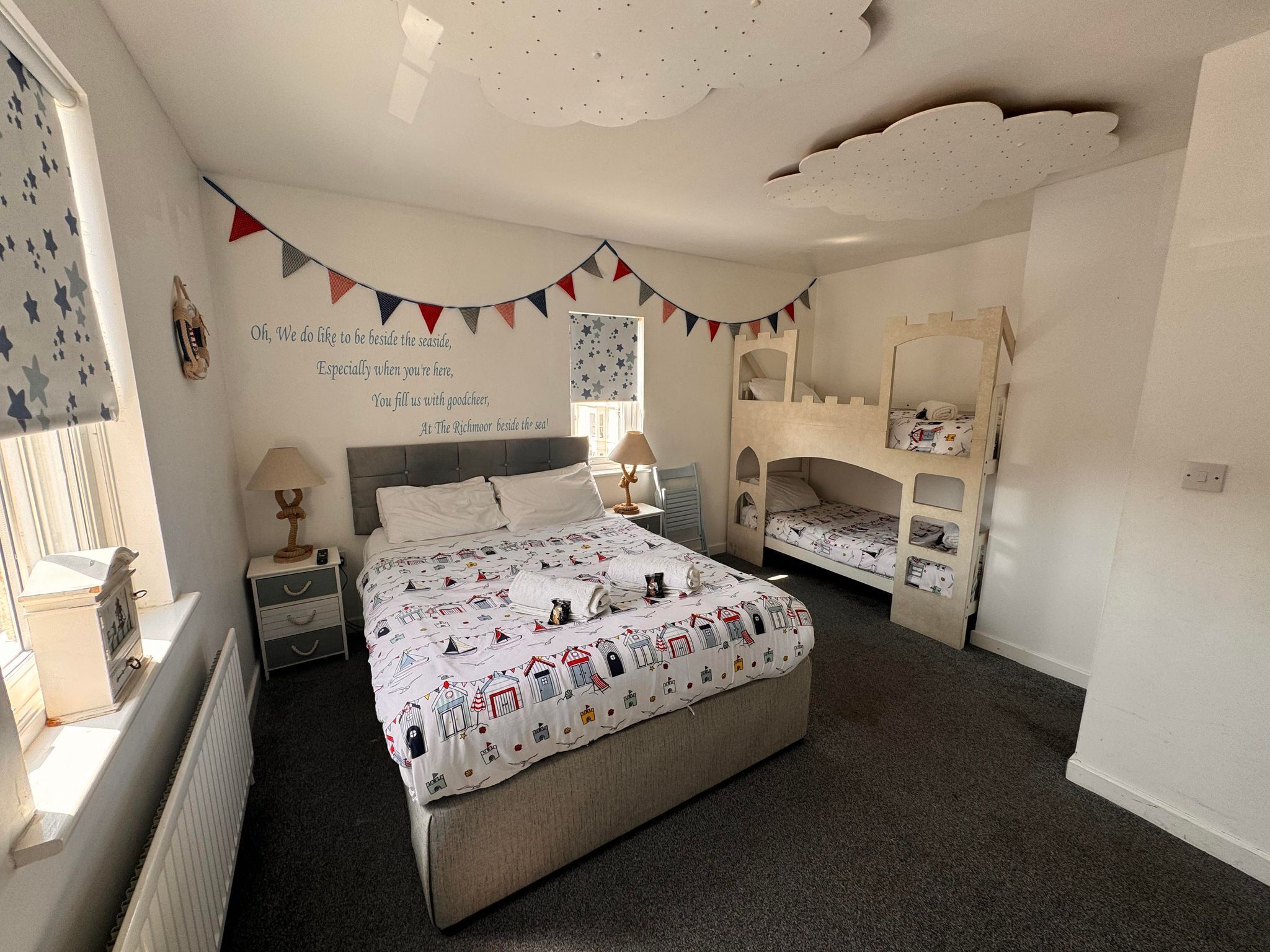 A bedroom with a bunk bed, a bed with a patterned duvet, a bedside table with a lamp, and a wall with a decorative banner and a poem.