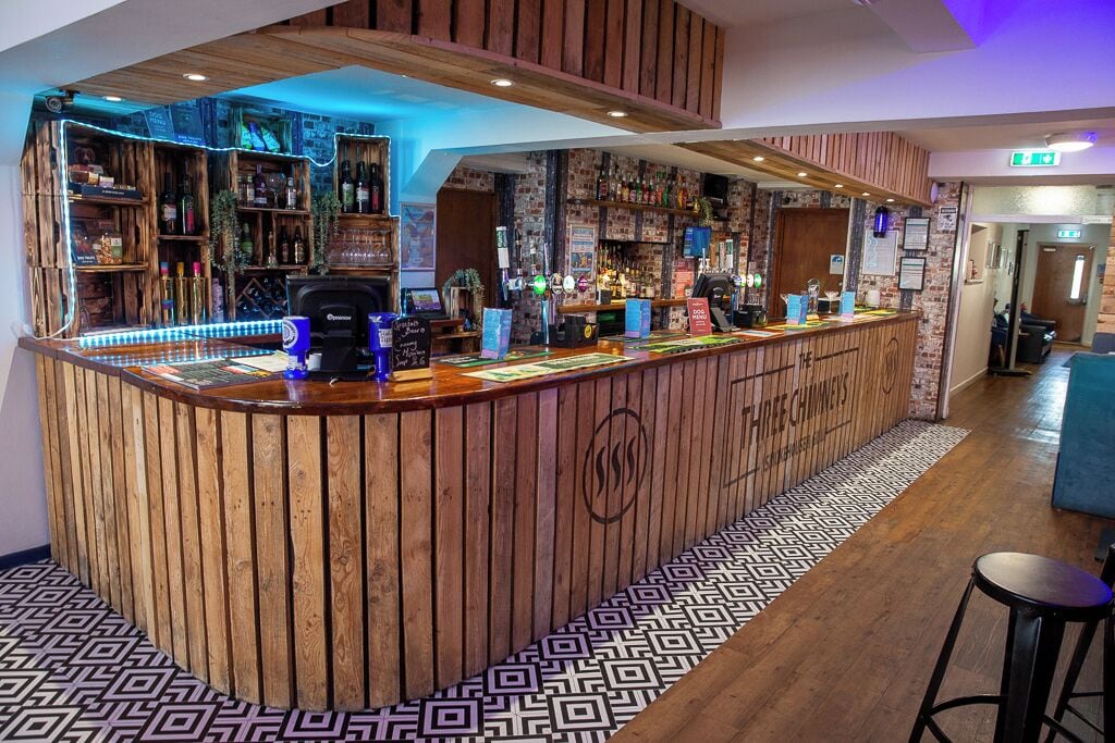 A bar area with a wooden counter, shelves with bottles, and a patterned floor.
