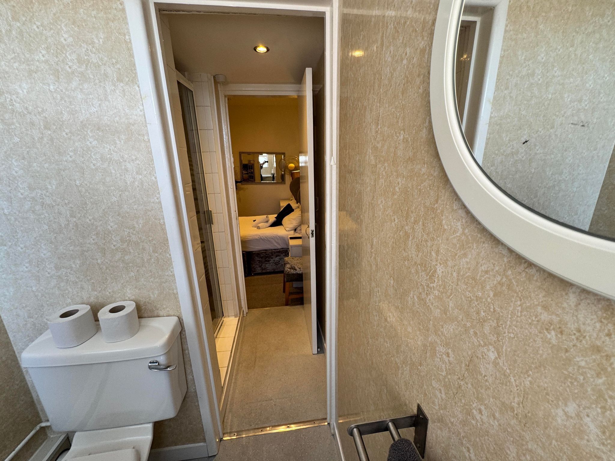 A narrow bathroom with a toilet, two rolls of toilet paper, a small bench, and a mirror.