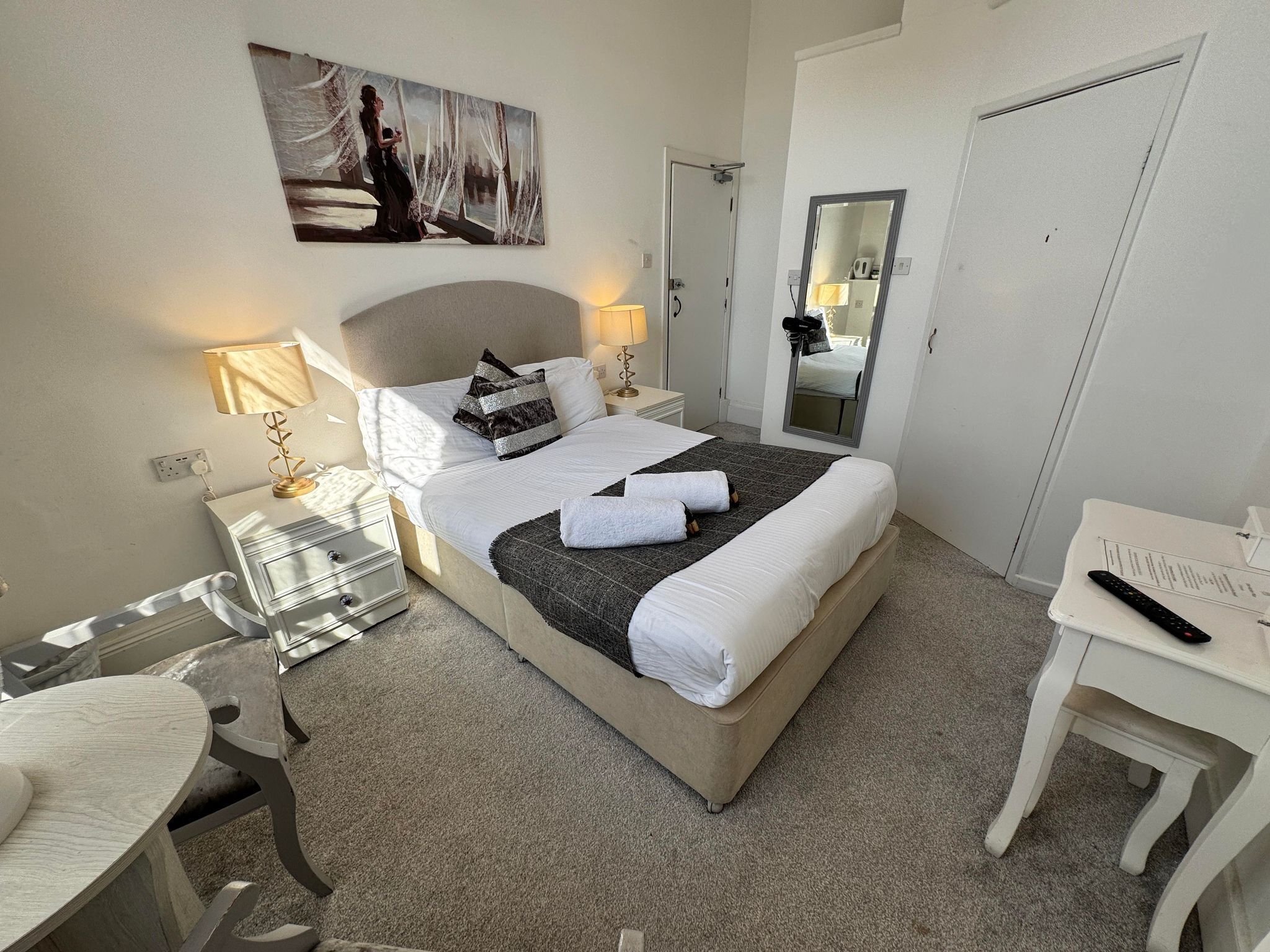 Habitación de hotel con cama, lámparas de noche, un escritorio, una silla, un espejo y un cuadro en la pared.