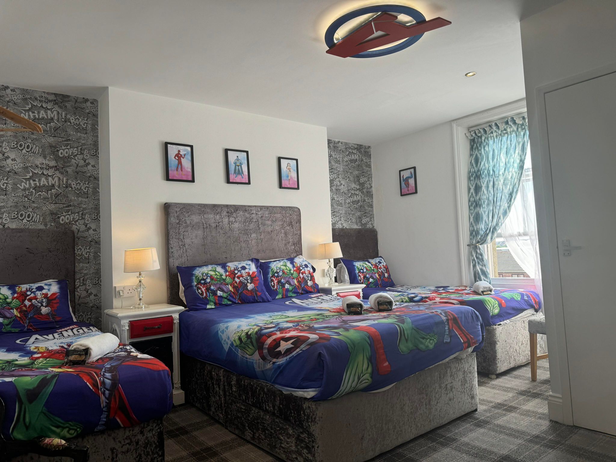 Habitación de hotel con dos camas, cada una con cubrecama con temática de superhéroe, cabeceros grises y una lámpara de techo.
