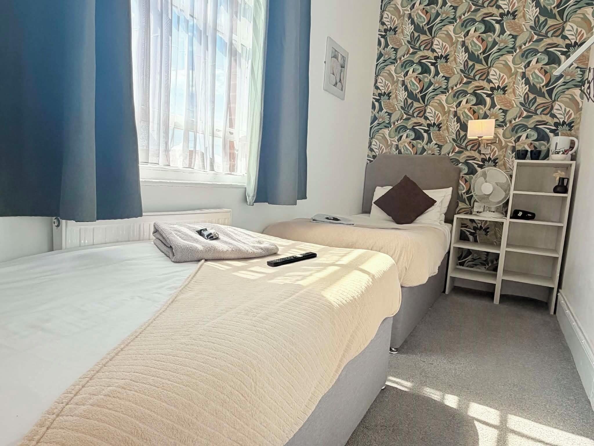 A hotel room with two beds, a window with curtains, a wall with a floral pattern, and a bedside table with a lamp.