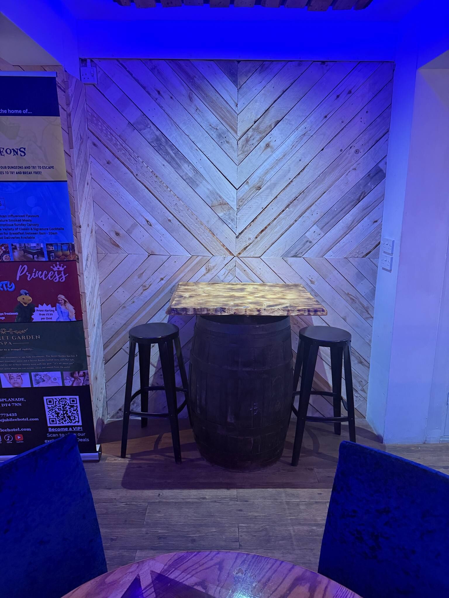 A wooden wall with a chevron pattern, a table with stools, and a sign with text and a QR code.