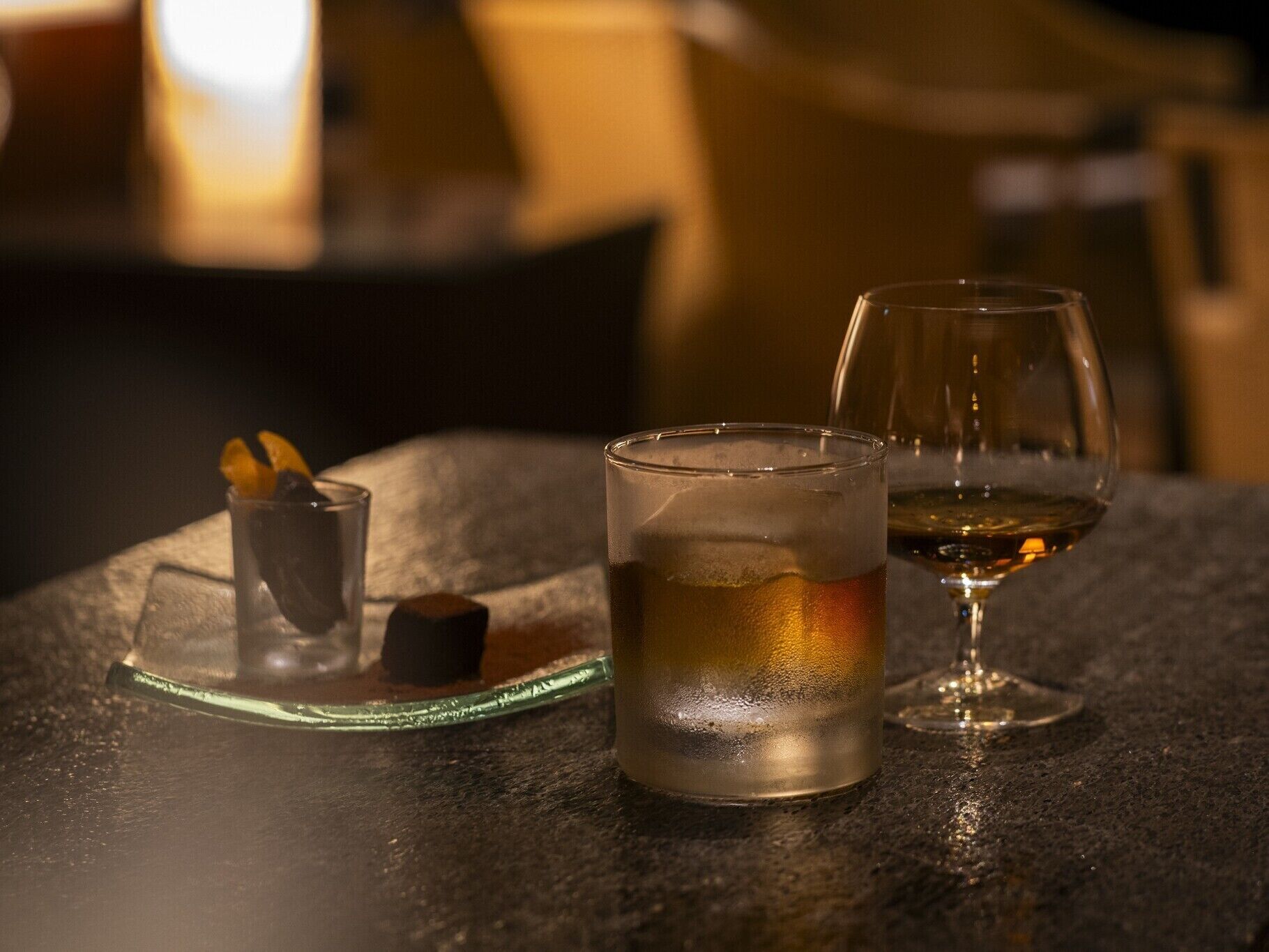 A table with a glass of amber liquid, a shot glass with ice and a dark cube, and an empty wine glass.