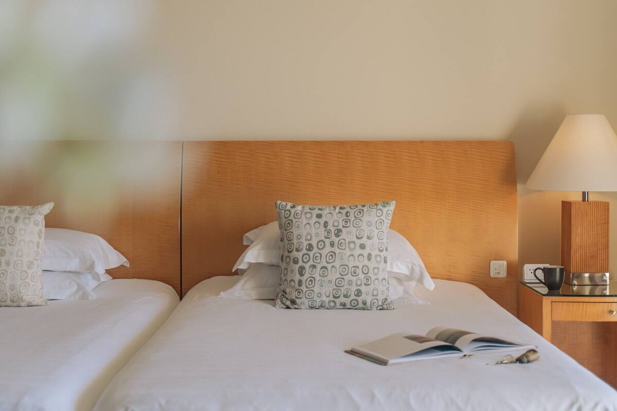 A hotel room with two beds, each with white linens and patterned pillows. There is a bedside table with a lamp, a cup, and a book.