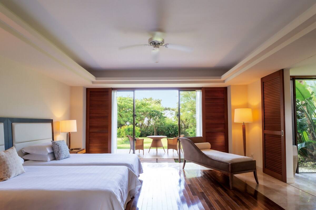 A hotel room with two beds, a ceiling fan, a dining area with chairs, and a view of greenery outside.