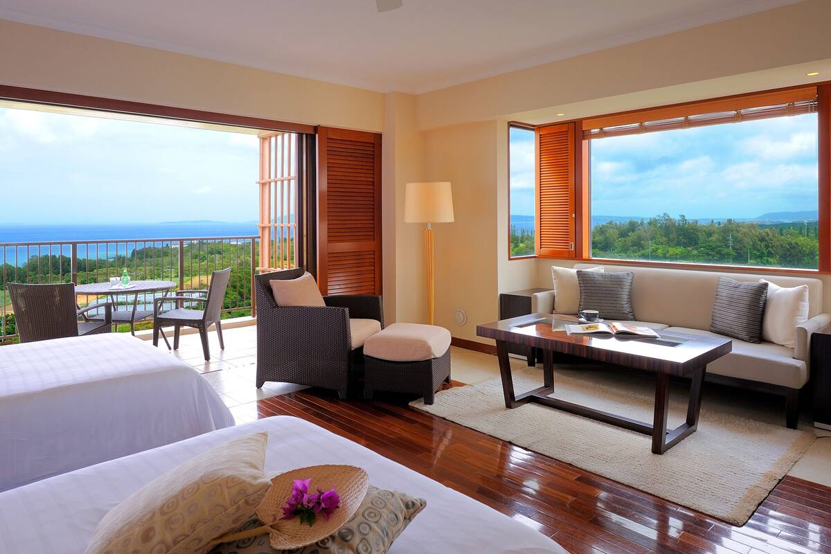 A hotel room with two beds, a sofa, a coffee table, and a balcony with a view of the ocean.