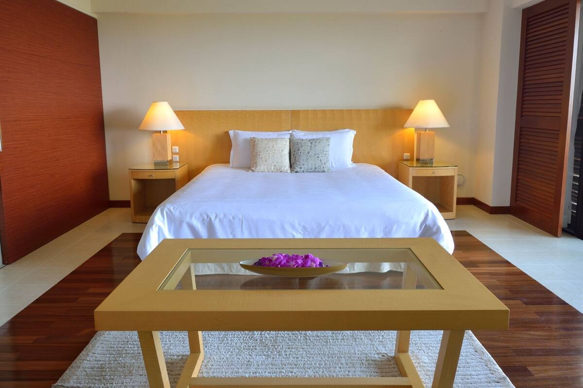 A hotel room with a large bed, two bedside lamps, a glass coffee table with a purple flower, and wooden flooring.