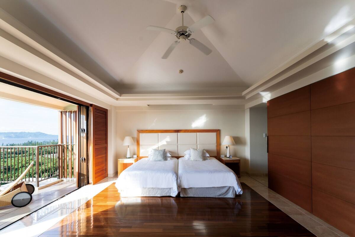 A spacious bedroom with a large bed, two bedside tables, a ceiling fan, and a balcony with a view.