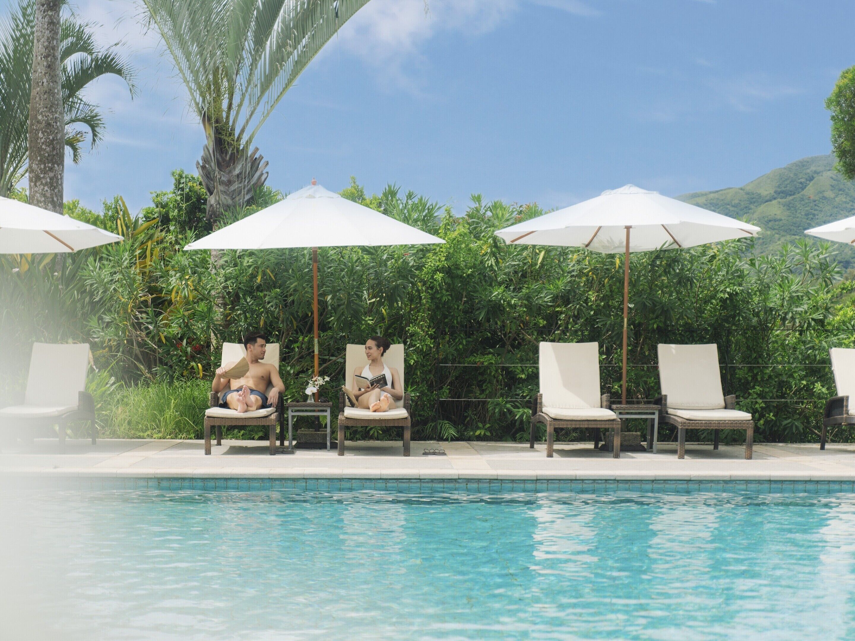 Un espace aménagé au bord de la piscine, avec des chaises longues et des parasols, entouré d’une végétation luxuriante et surplombé d’un ciel dégagé.