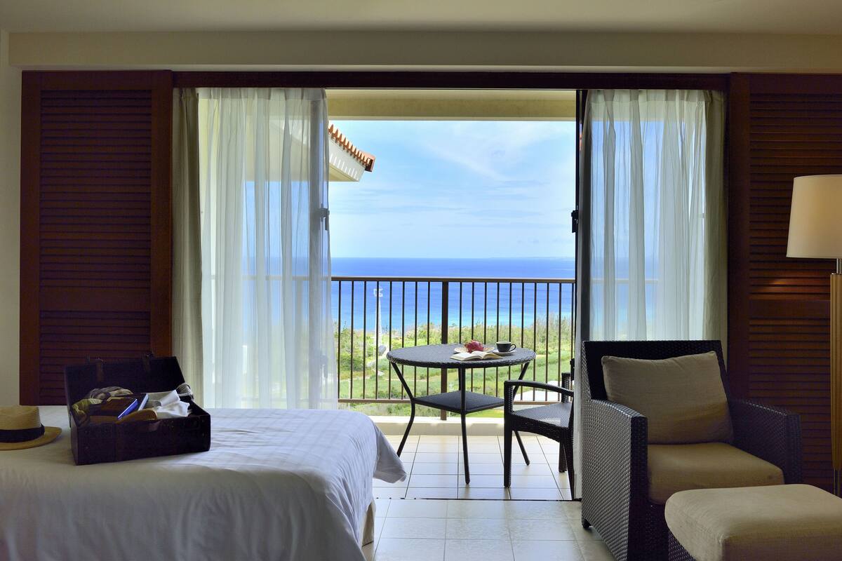 A hotel room with a bed, a small table with a cup and book, a chair, and a view of the ocean through a balcony.