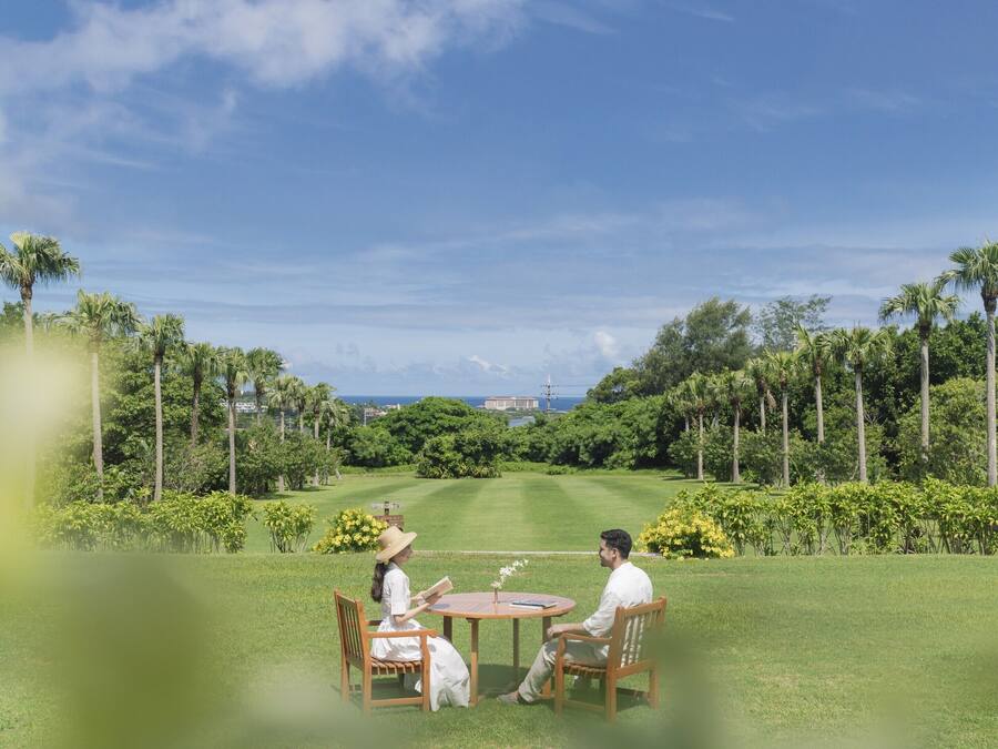 A couple sitting at a round wooden table in a lush garden with palm trees and a view of the ocean.