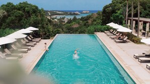 A rectangular pool with clear blue water, surrounded by lounge chairs and umbrellas, with a view of the sea and buildings in the background. - The Atta Terrace Club Towers (Onna)