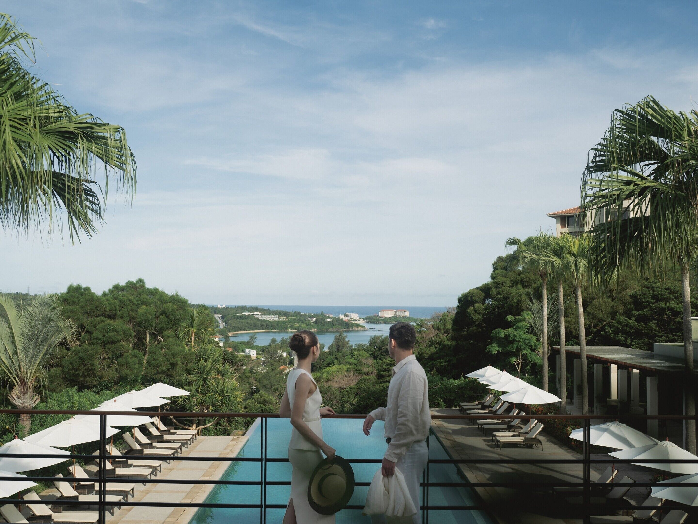 A couple overlooking a pool with lounge chairs and umbrellas, surrounded by lush greenery and a view of a distant town.
