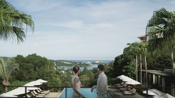 A couple overlooking a pool with lounge chairs and umbrellas, surrounded by lush greenery and a view of a distant town.