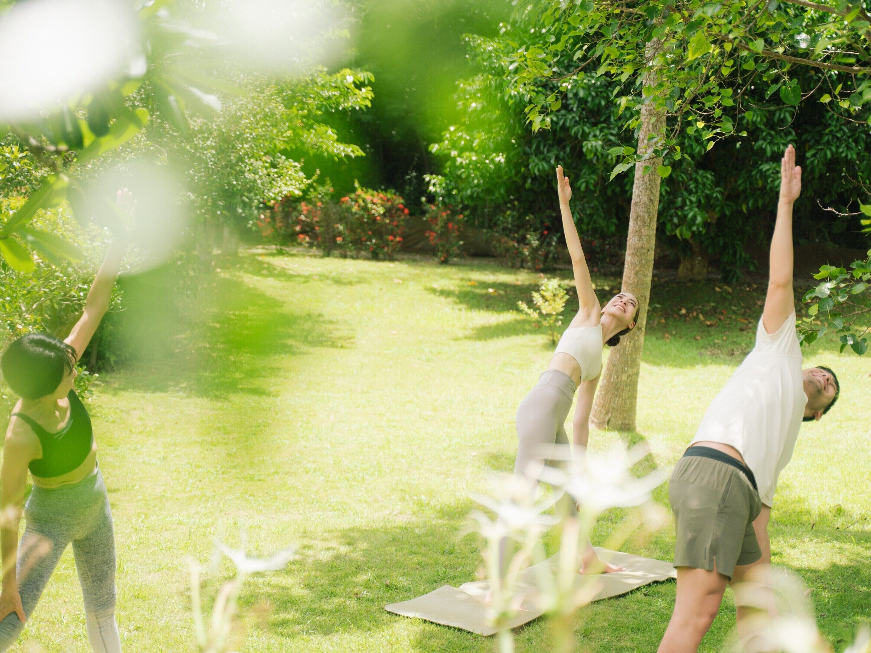 A group of people practicing yoga outdoors on a grassy area with trees and flowers in the background.