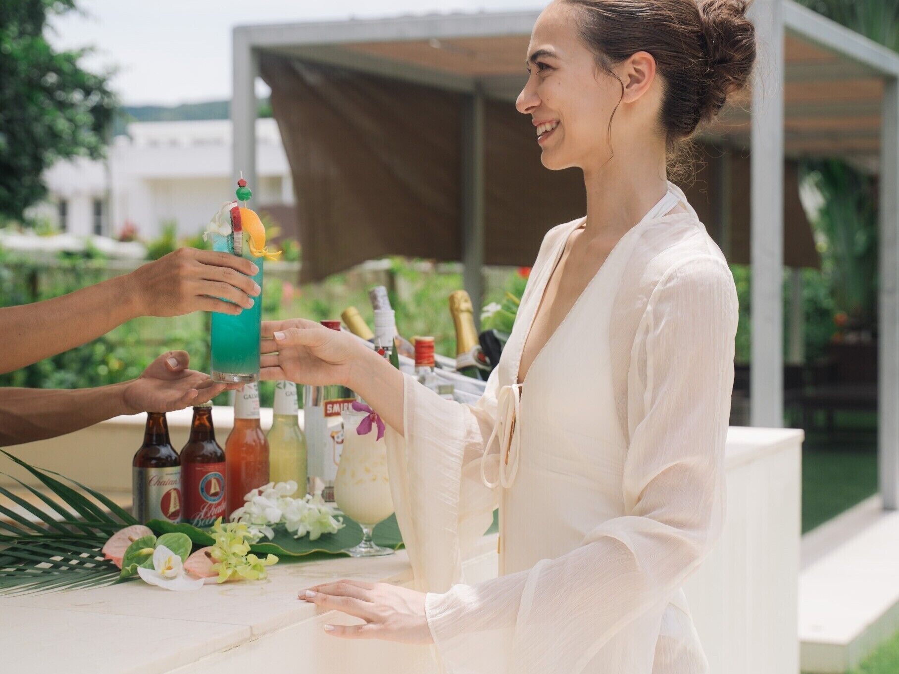 Iemand houdt een cocktail vast aan een bar met diverse flessen en een glazen kan.