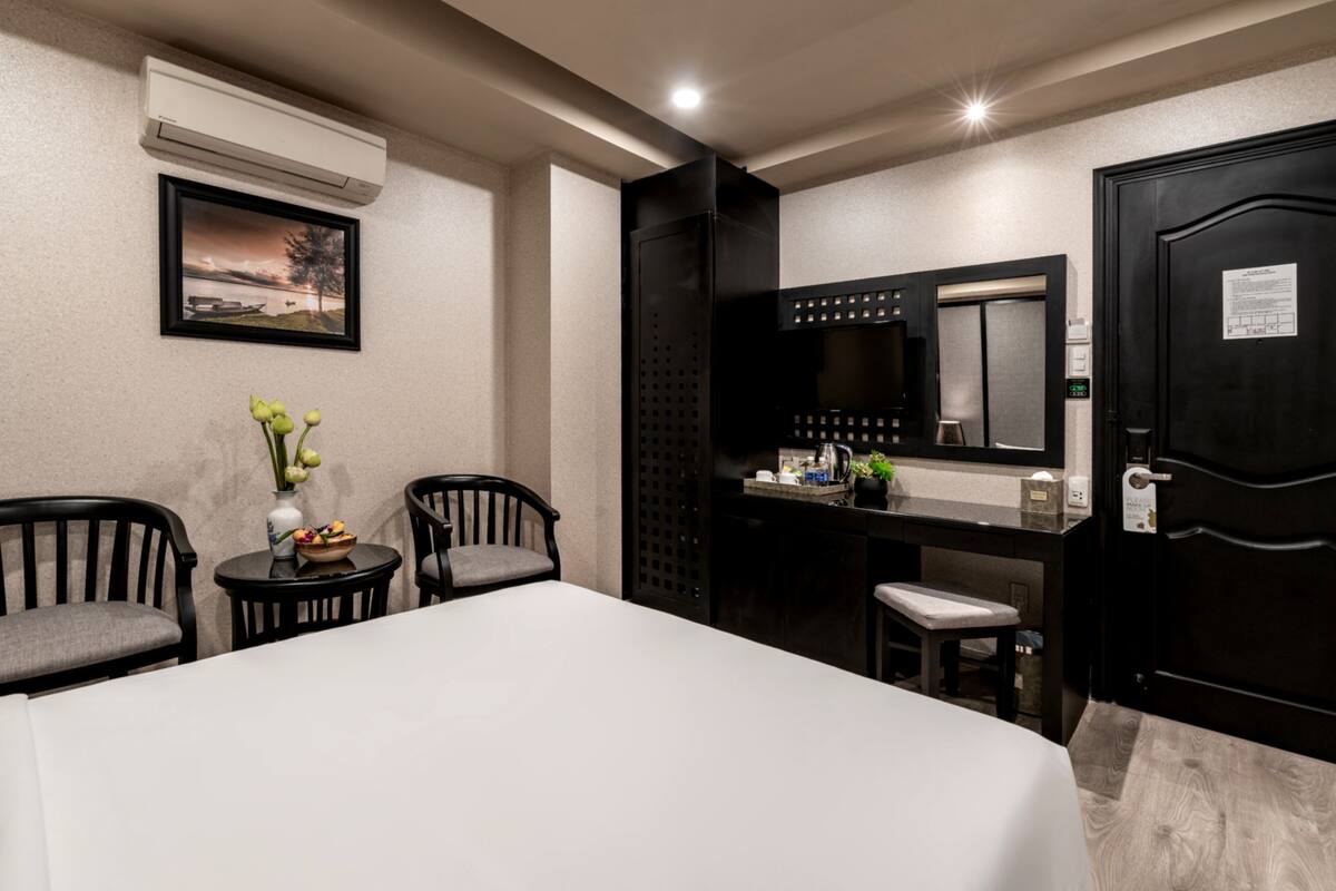 A hotel room with a bed, two chairs, a small table with a vase of flowers, a mirror, and an air conditioning unit on the wall.