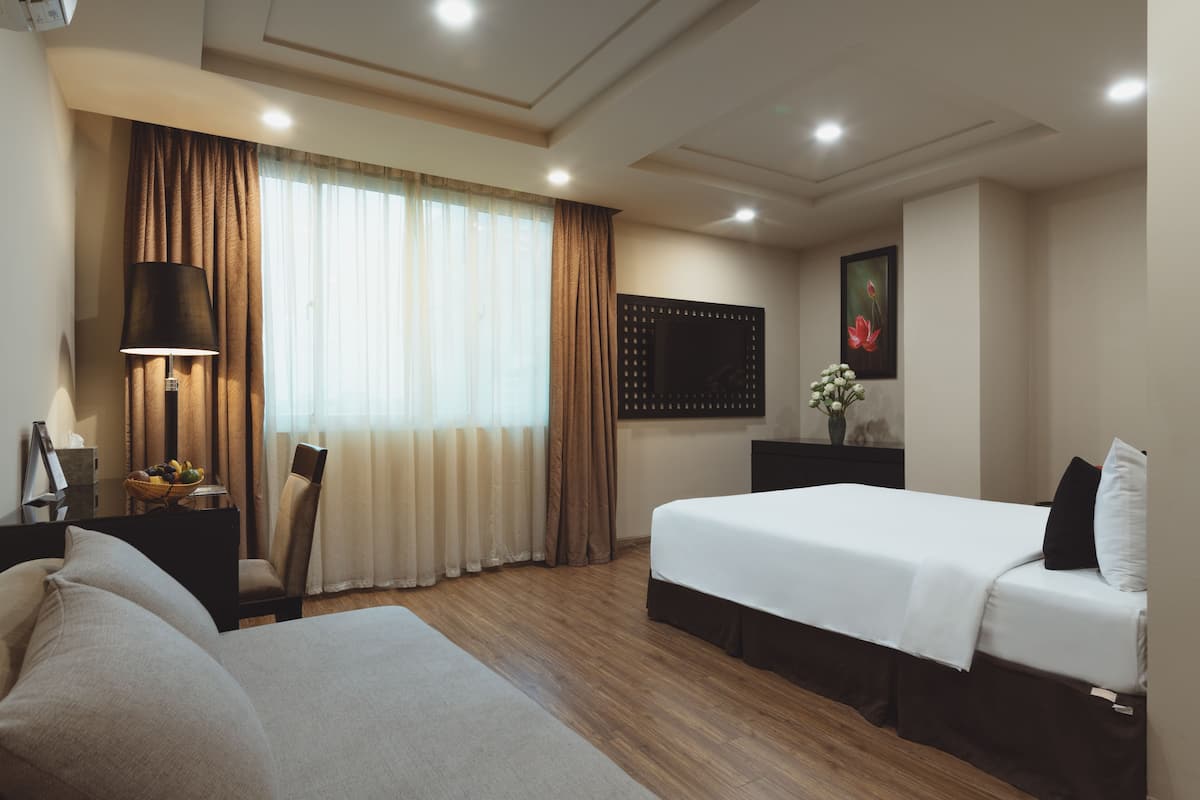 A hotel room with a bed, a desk with a lamp, a chair, a large window with curtains, and a wall-mounted TV.