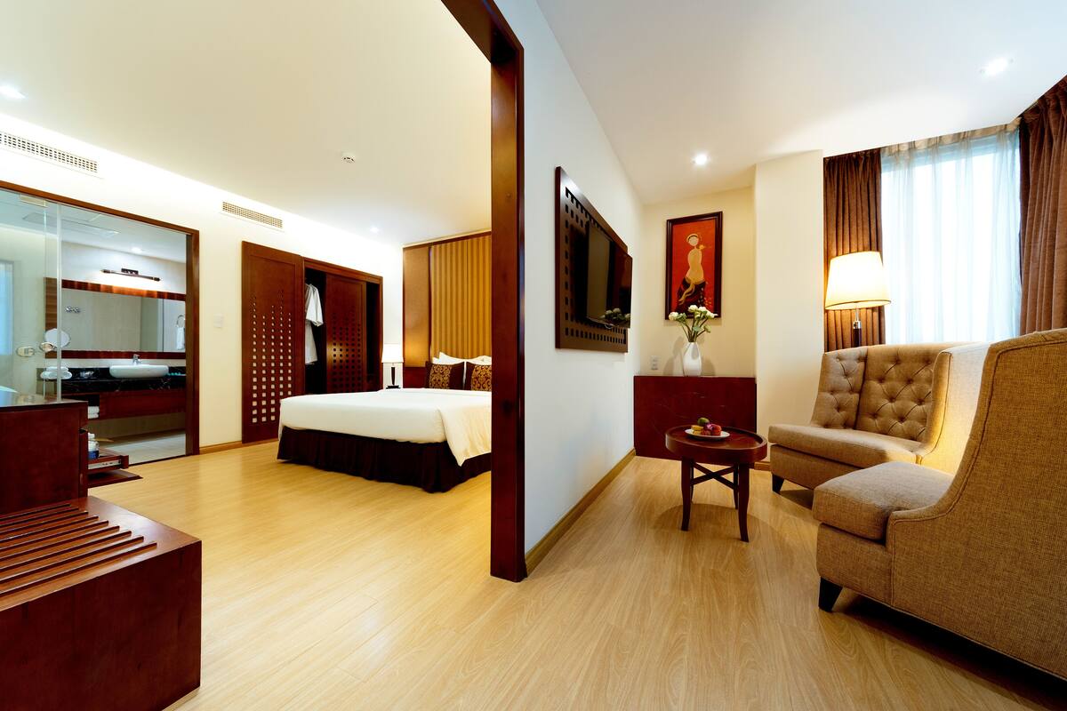 A hotel room with a bed, a sofa, a small table, a mirror, a TV, and a bathroom with a sink and mirror.