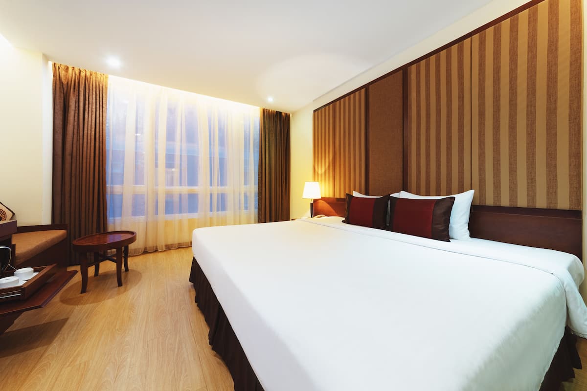 A hotel room with a large bed, a desk with a chair, a lamp, and a view of the city through the window.