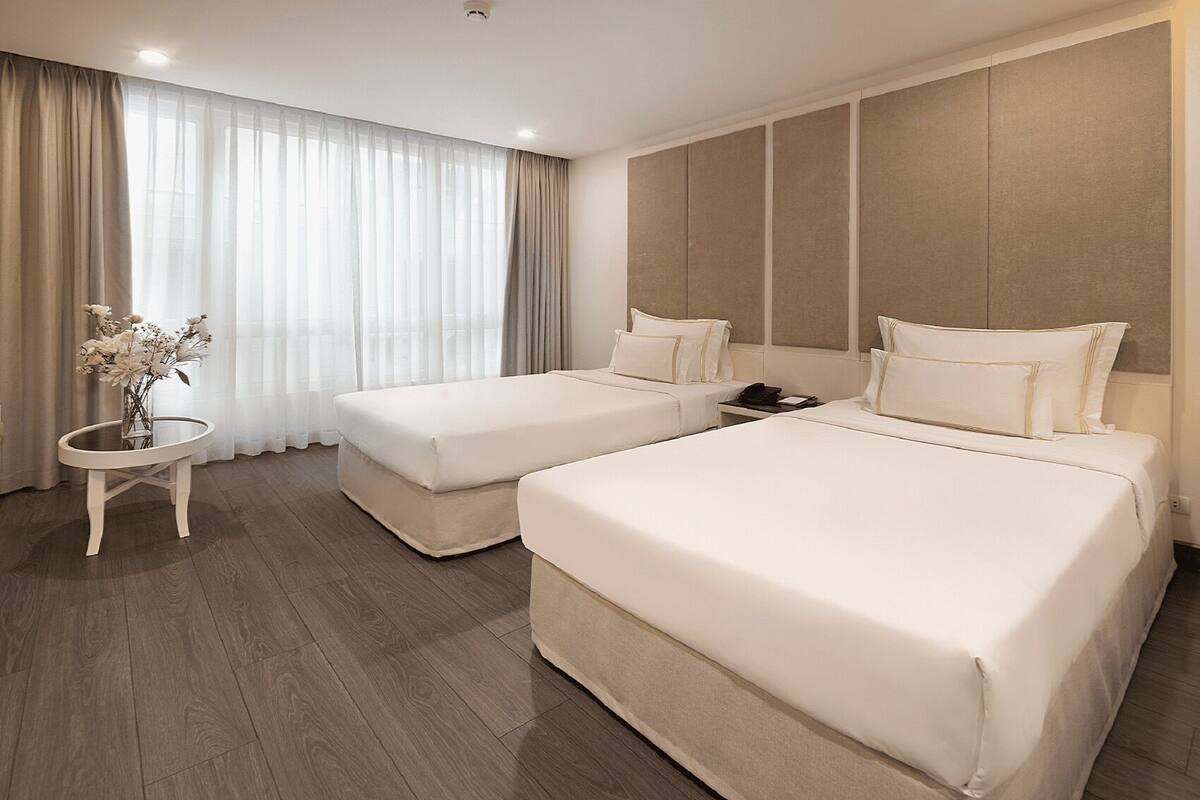 A hotel room with two single beds and one double bed, a small round table with a vase of flowers, and a large window with sheer curtains.
