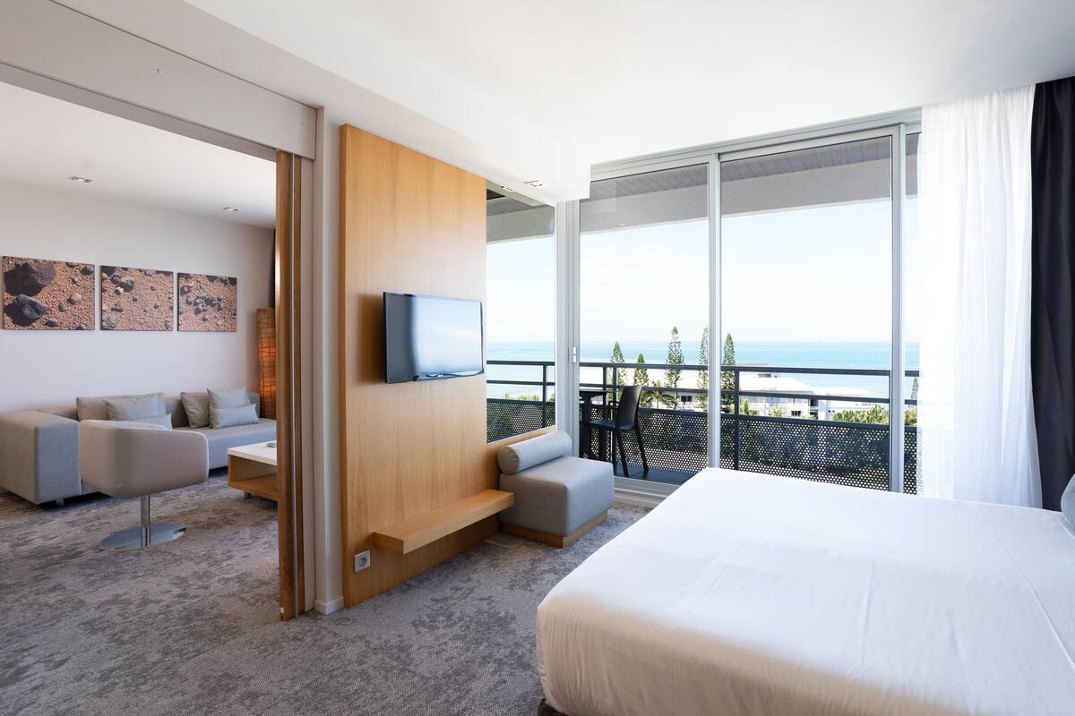 Quarto de hotel moderno com uma cama grande, um sofá, uma televisão e uma varanda com vista para o mar.