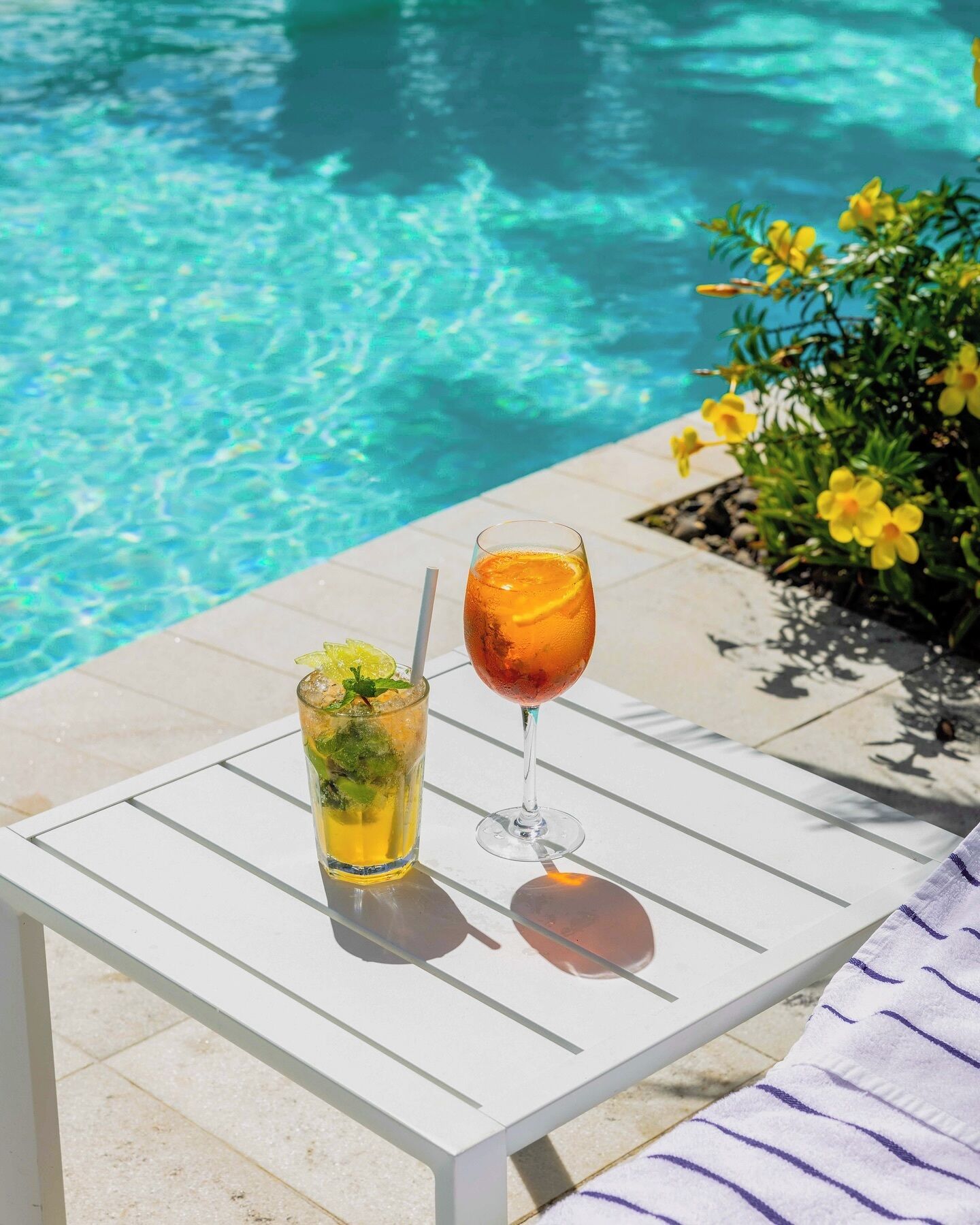 A poolside table with two drinks and a pool in the background.