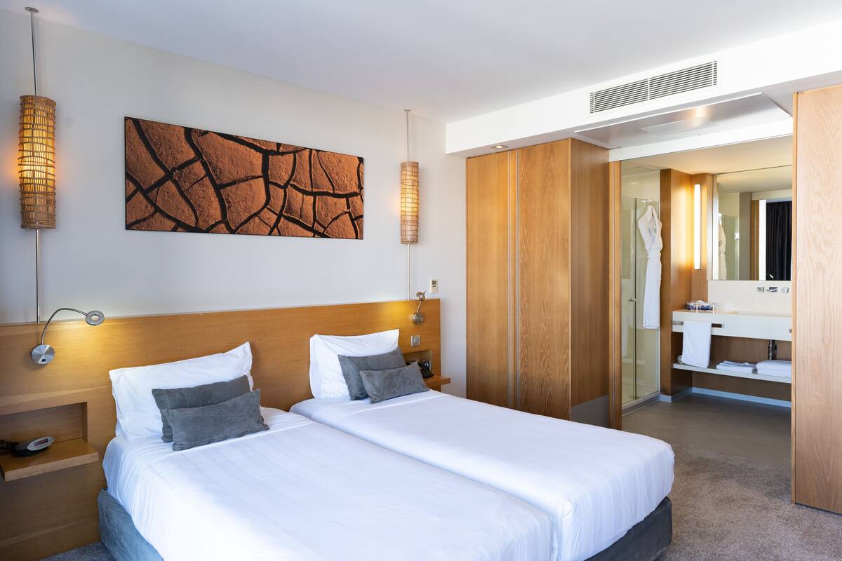 Quarto de hotel moderno com uma cama grande, cabeceira de madeira, uma obra de arte na parede e um banheiro visível por uma porta aberta.