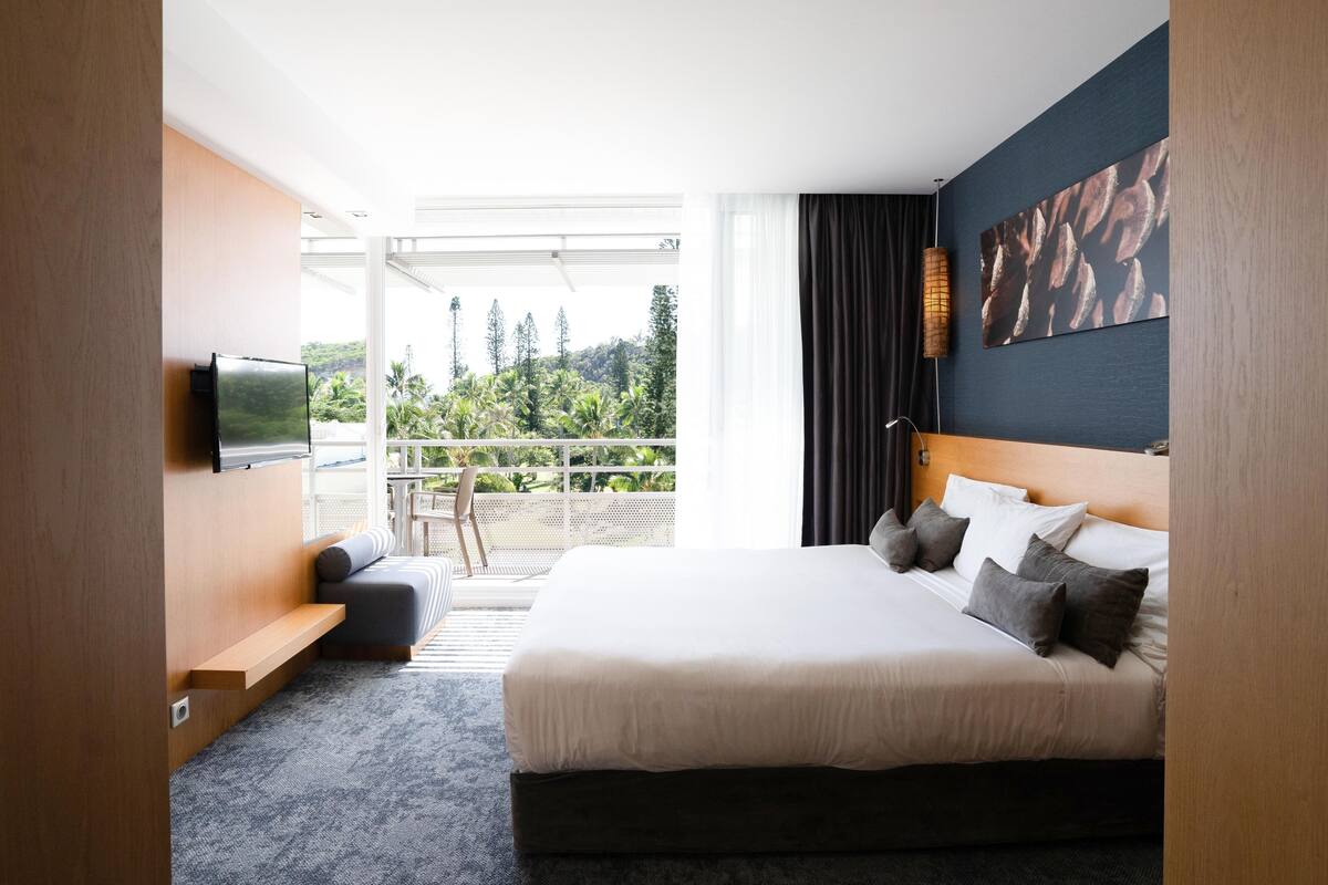 Quarto de hotel moderno com uma cama grande, televisão, varanda com vista para áreas verdes e uma parede de destaque em madeira.