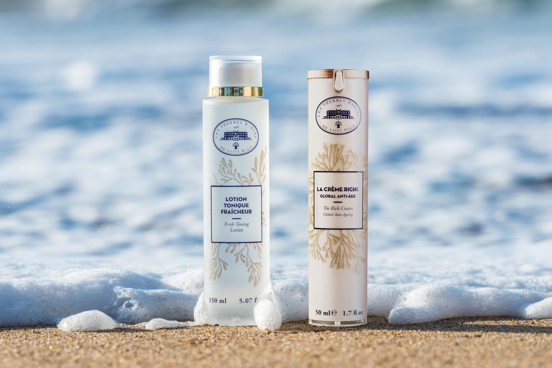 Two bottles of lotion on a sandy beach with waves in the background.