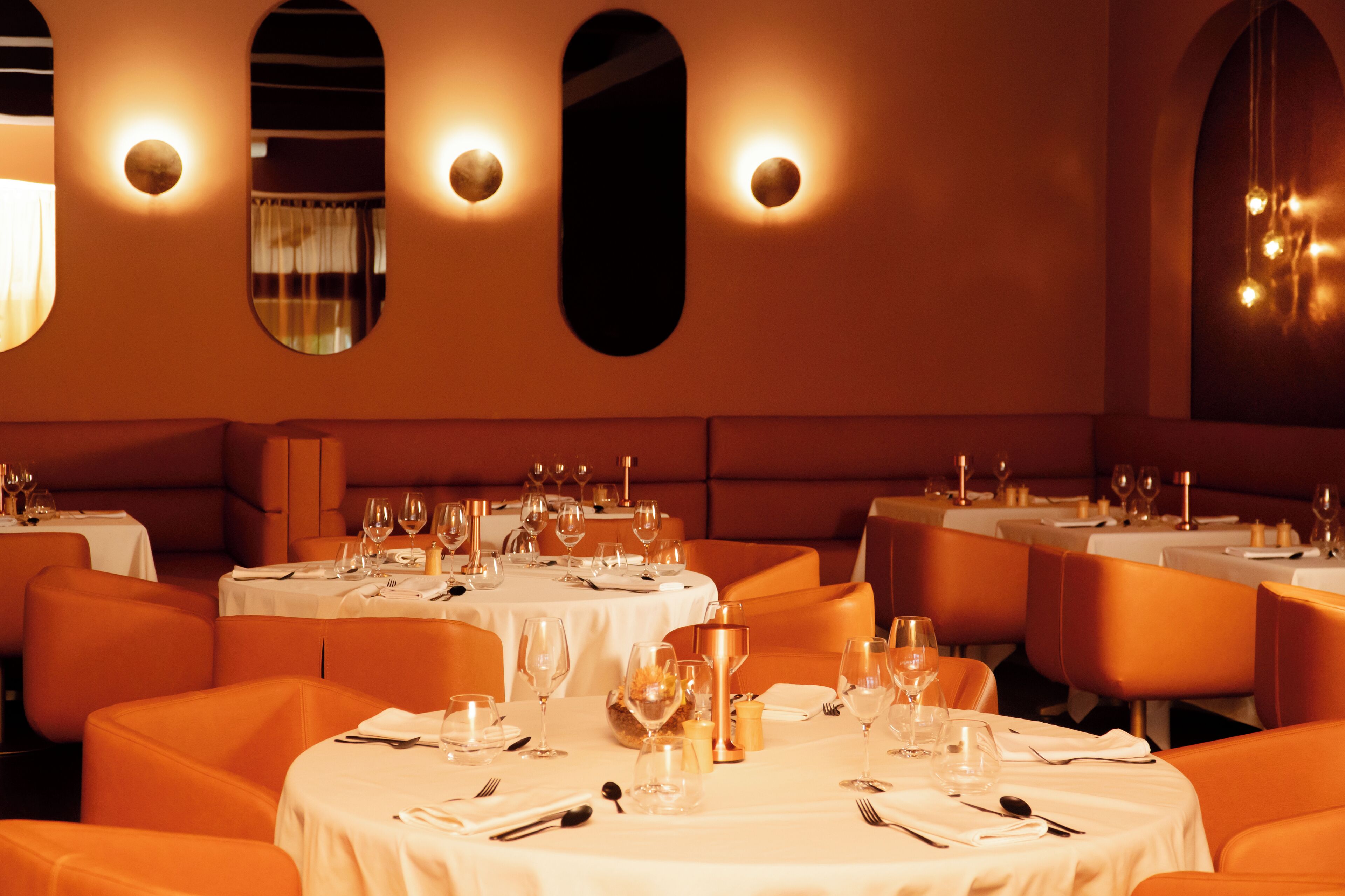 A restaurant with round tables set for dining, orange seating, and wall sconces.