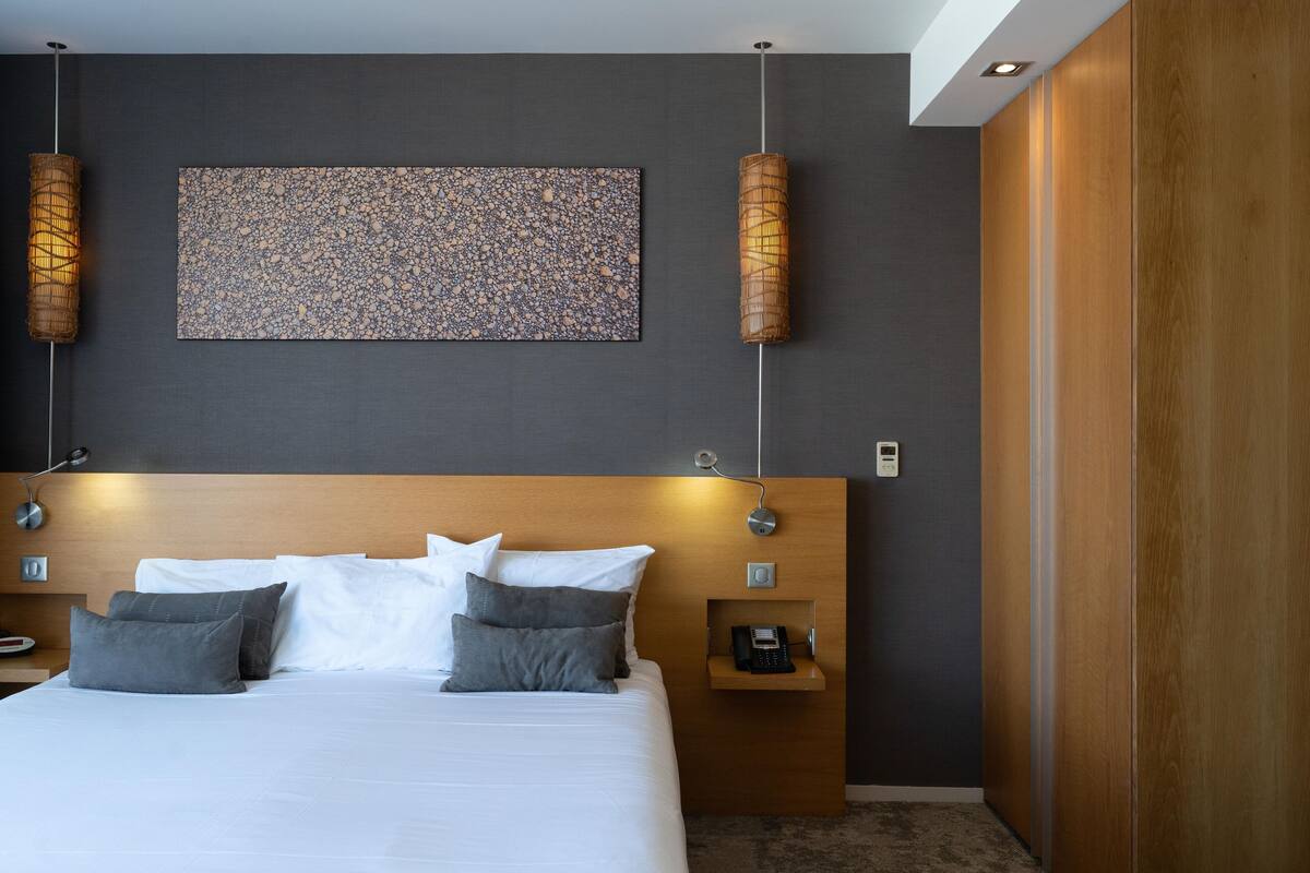 Quarto de hotel moderno com uma cama grande, cabeceira de madeira, uma obra de arte na parede e duas luminárias de parede.