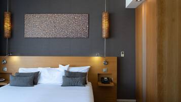 A modern hotel room with a large bed, a wooden headboard, a wall-mounted artwork, and two wall-mounted lamps.