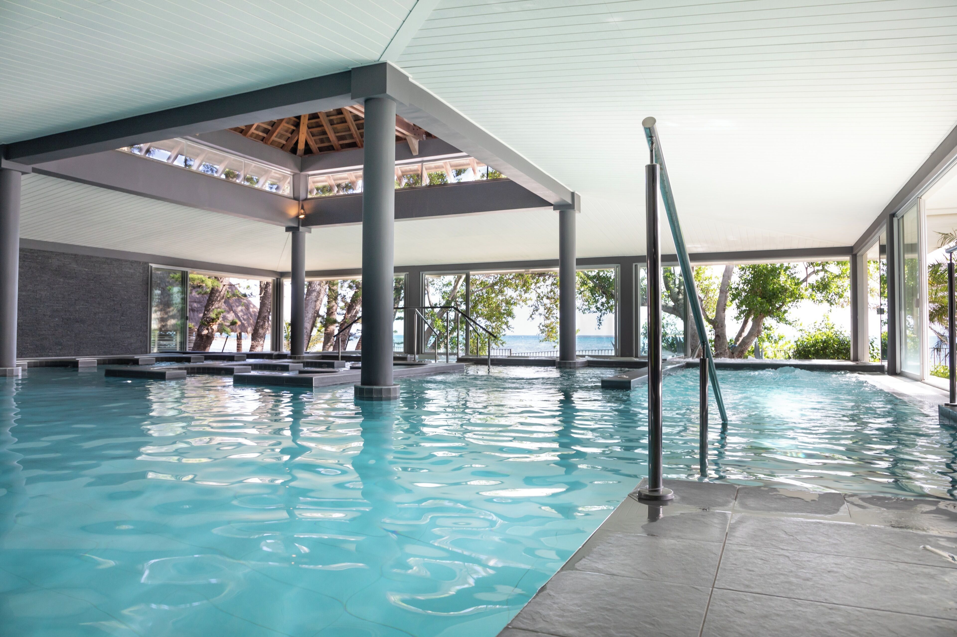 Una piscina interior con agua cristalina, una zona cubierta con pilares y grandes ventanales que ofrecen vistas a árboles y una masa de agua.