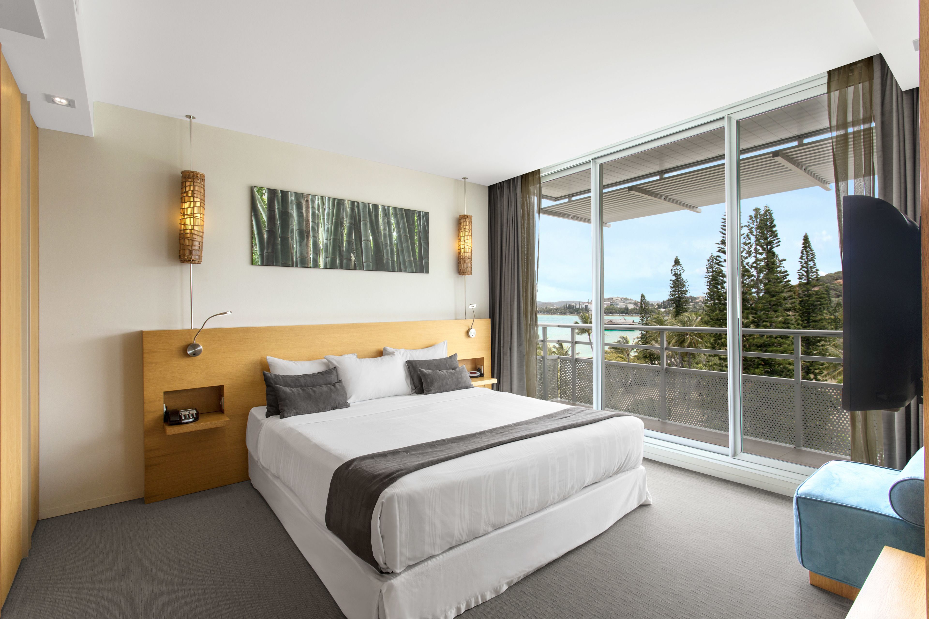 A modern hotel room with a large bed, a wooden headboard, a view of trees and mountains through floor-to-ceiling windows, and a balcony with a view.