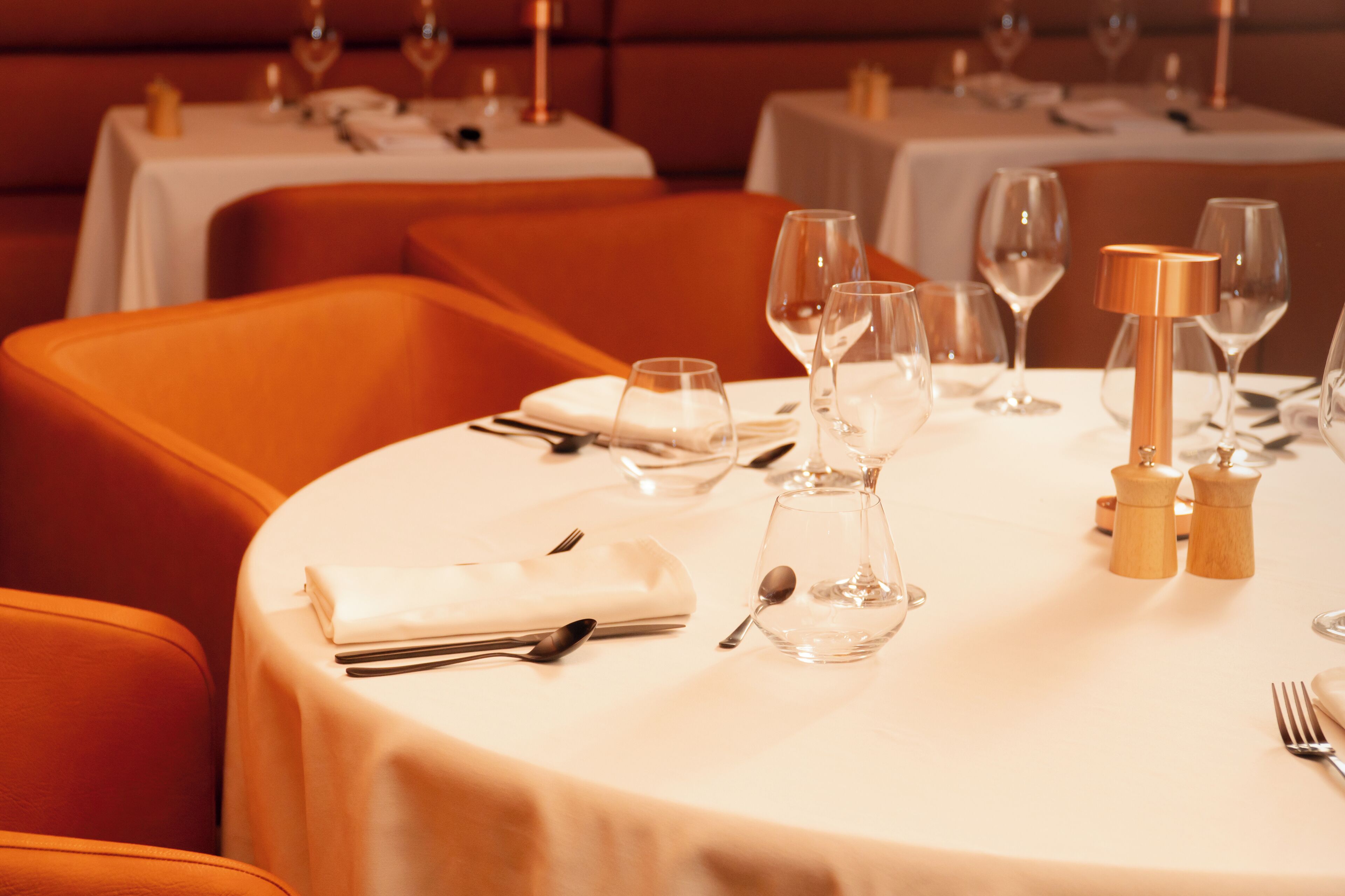 A dining table set with white tablecloth, silverware, and wine glasses, with orange upholstered chairs and a white table in the background.