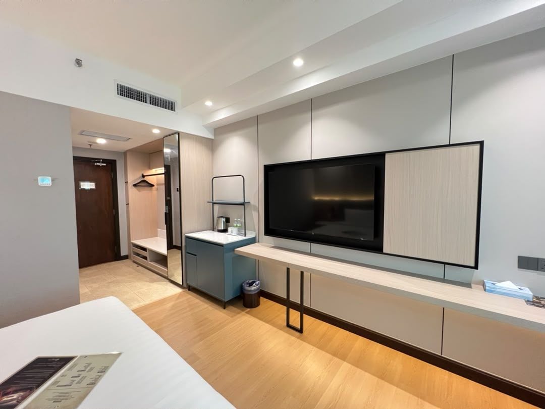 A modern hotel room with a large flat-screen TV mounted on the wall, a small kitchenette area, and a wooden floor.