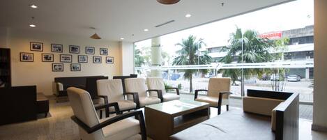 A hotel lobby with a reception desk, seating area, and large windows offering a view of the street and palm trees.