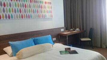 A hotel room with a bed, blue and white pillows, a desk with a chair, a wall with a leaf pattern, and a window with curtains.
