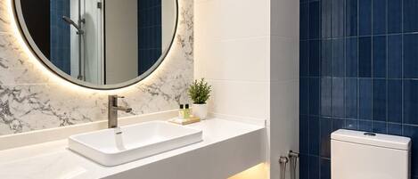 A modern bathroom with a marble vanity, a round mirror with underglow, a toilet, and blue-tiled shower area.