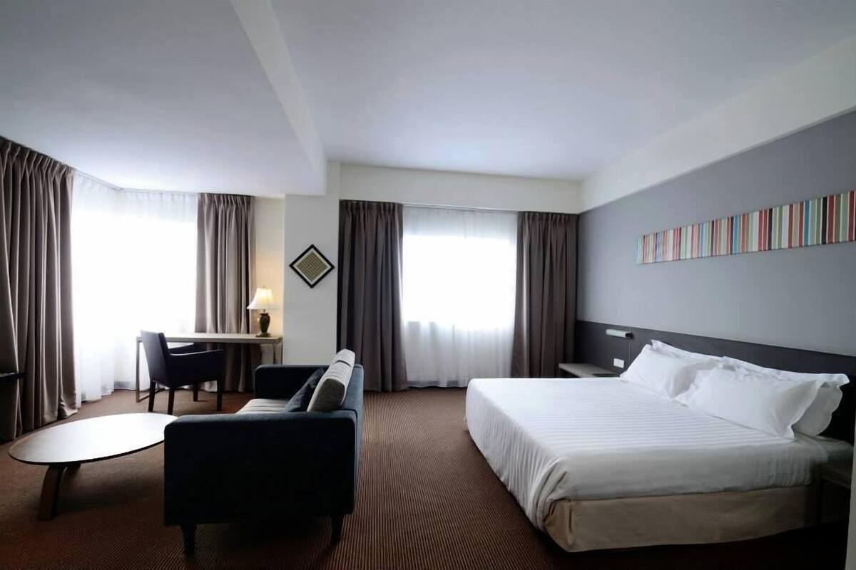 A hotel room with a large bed, a desk with a chair, a small table, and a wall with a striped artwork.