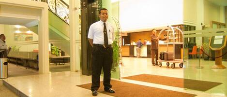A man standing in a hotel lobby with a reception desk, a luggage cart, and a staircase in the background.
