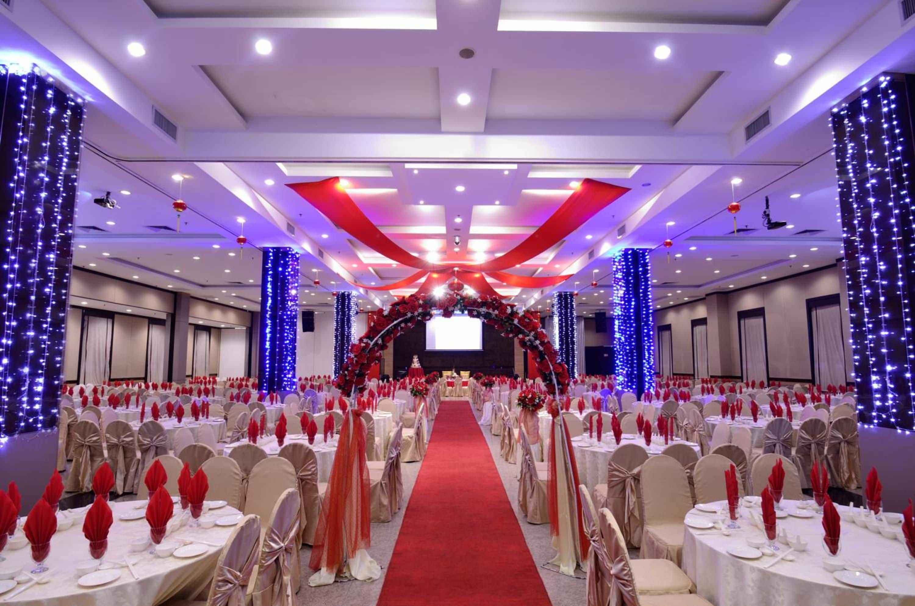 A banquet hall with round tables, red and white table settings, and a red carpet leading to a stage.