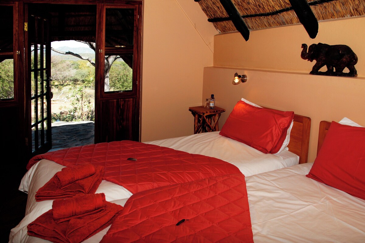 A room with two beds, red bedding, a small table with a bottle, a wall-mounted elephant sculpture, and a view of the outdoors through an open door.
