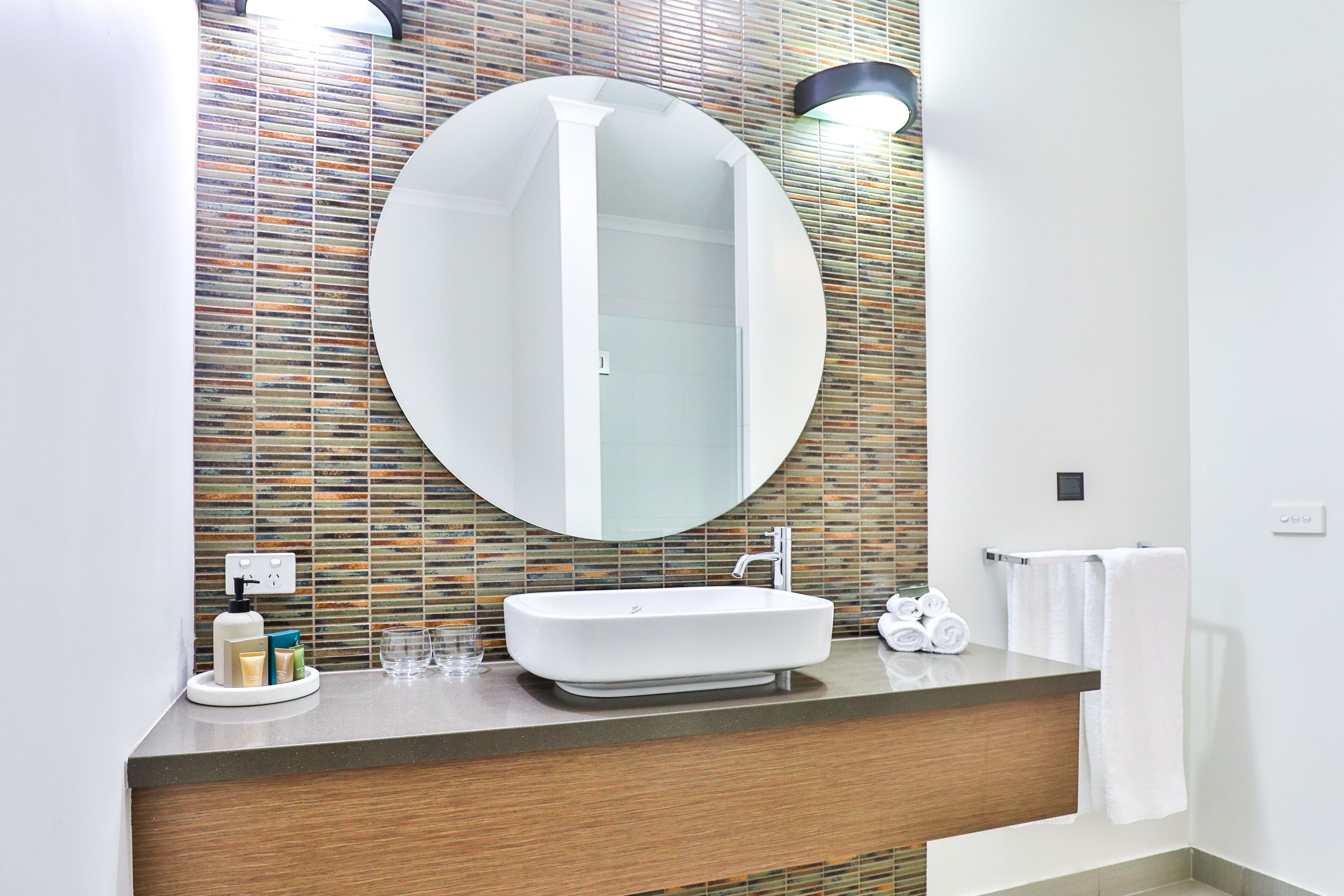 A modern bathroom with a rectangular sink, a large round mirror, and a tiled backsplash.