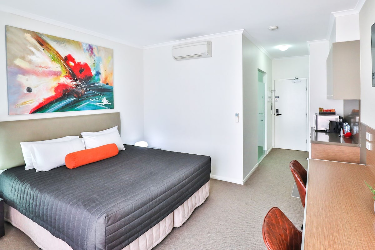 Une chambre d’hôtel avec un lit, un bureau, une chaise, une télévision, un tableau et une salle de bain visible à travers une porte ouverte.